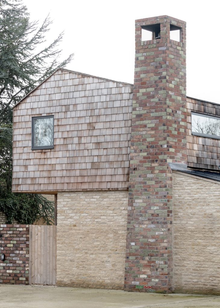 A rural new-build house featuring a tall, multi-coloured brick chimney beside a wall of light-coloured stone, with wooden shingles cladding the upper storey and two windows.