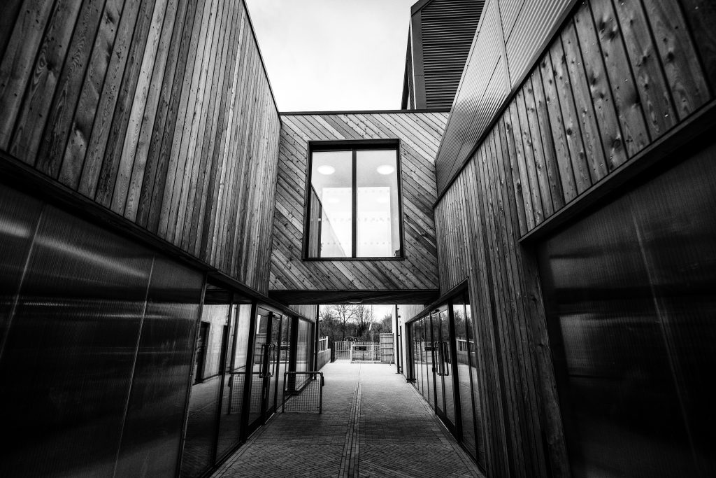 Black and white photo of a modern building corridor with wood panelling highlights sleek community architecture. Glass doors line both sides, while a rectangular window above the walkway frames the cloudy sky in this geometric perspective.