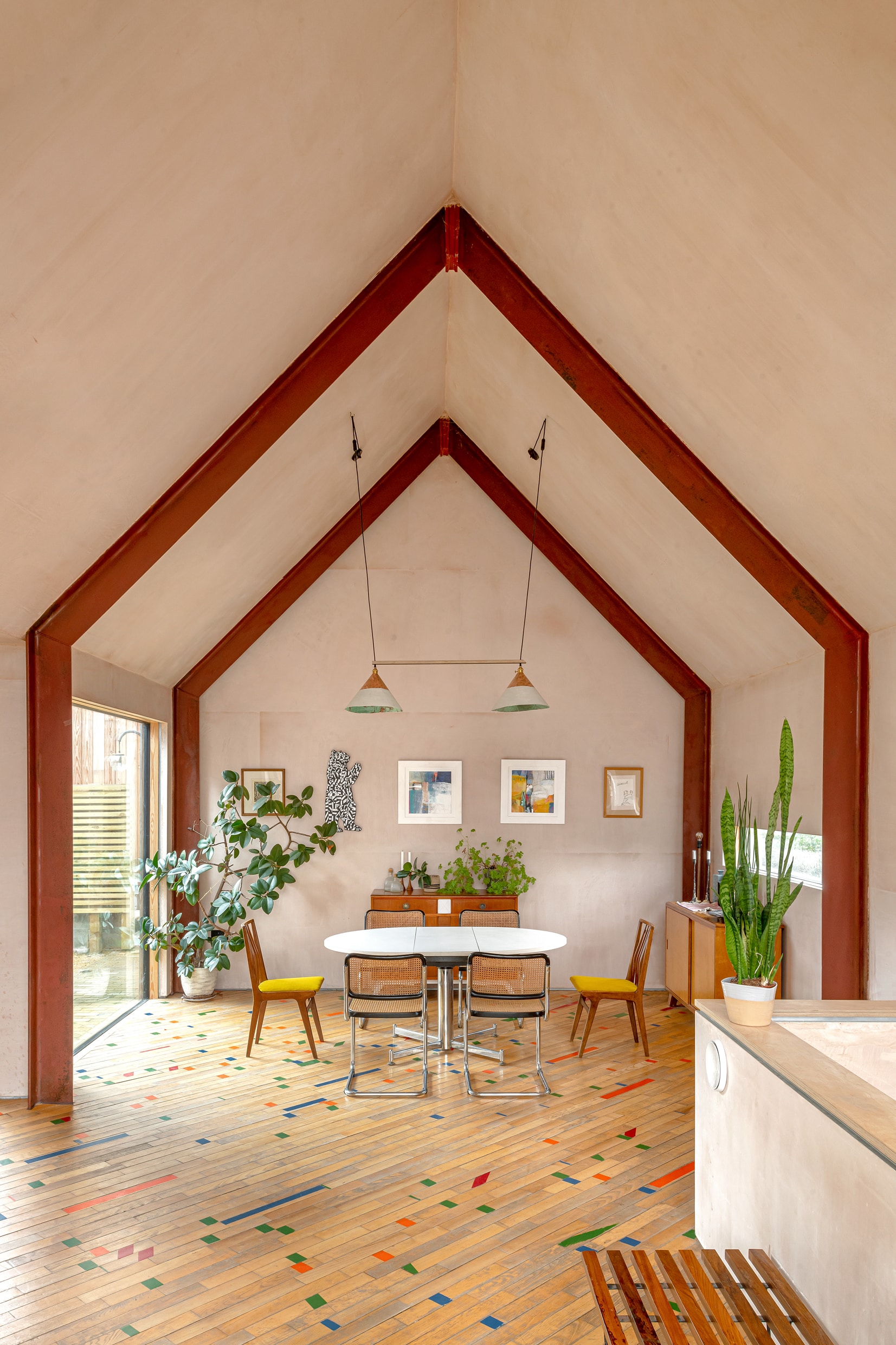 A bright dining area in this Eco New Build House features a high vaulted ceiling with exposed red beams, wooden floors with colourful tile accents, mismatched chairs around a wooden table, green plants, framed art, and large windows for natural light.
