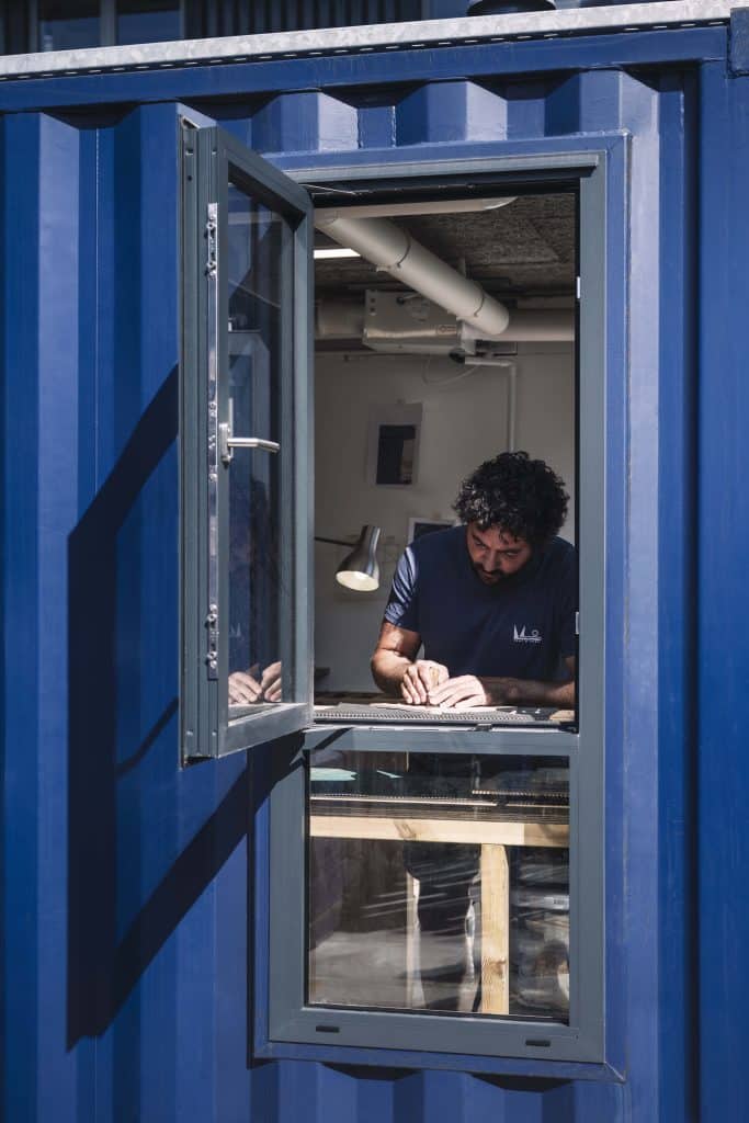 A person working at a desk inside a blue container-like structure, visible through an open window. The desk is cluttered with papers and tools, and the interior has industrial features such as exposed pipes and lighting fixtures