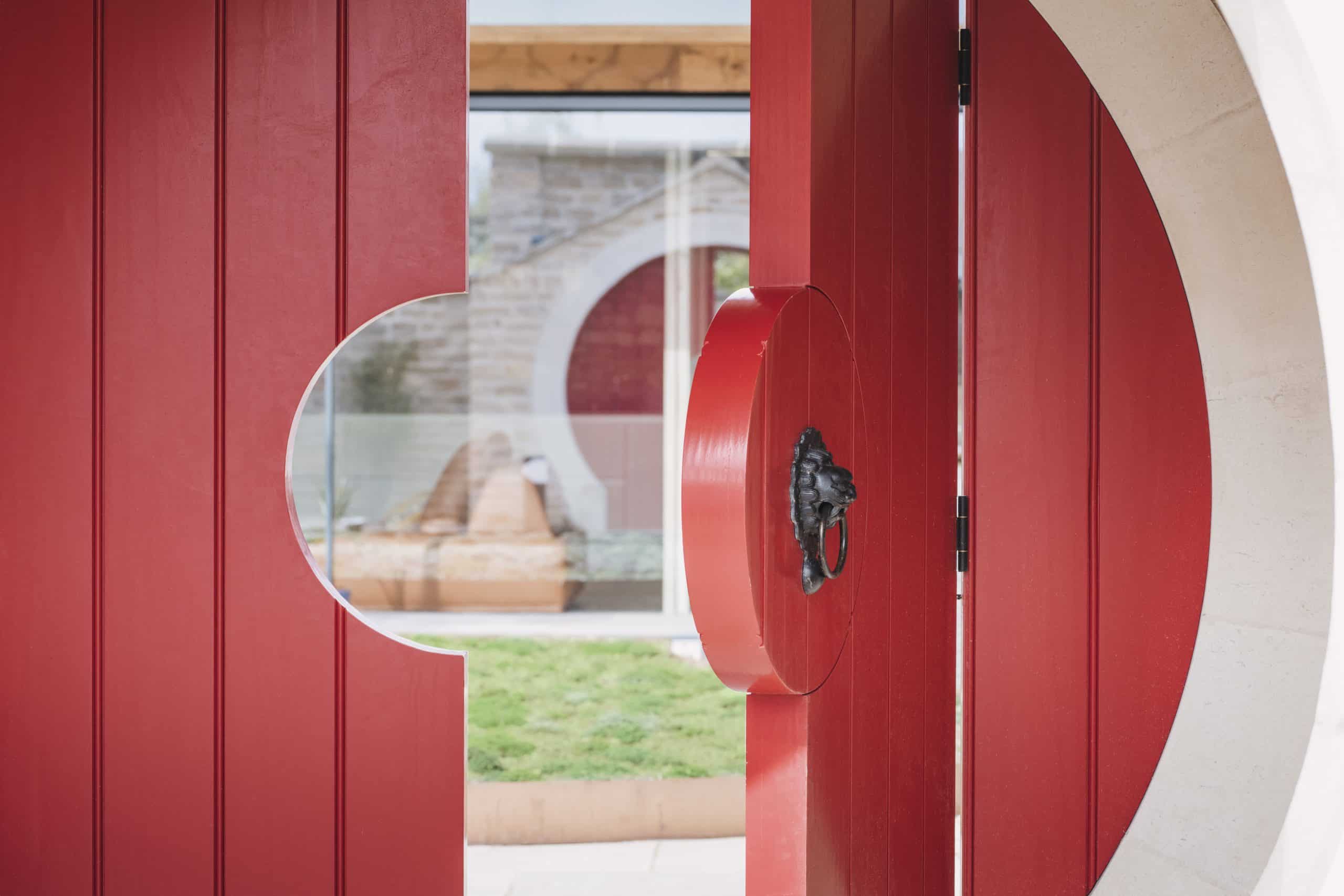 A bold red circular door, partially open, features a cut-out semicircle and a metal lion head handle. Through the doorway of this listed barn conversion in the Cotswolds, a glass wall and another red circle reveal grass and a stone structure outside.