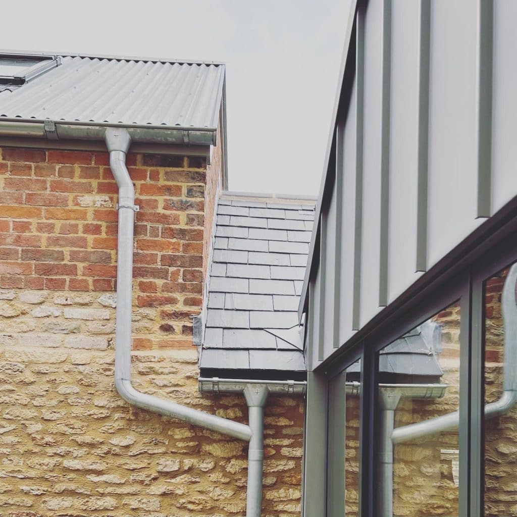 A close-up of two adjoining buildings: one with a stone wall and tiled roof with metal gutters, and the other with a modern facade of vertical metal panels and large reflective windows. The sky above is overcast.