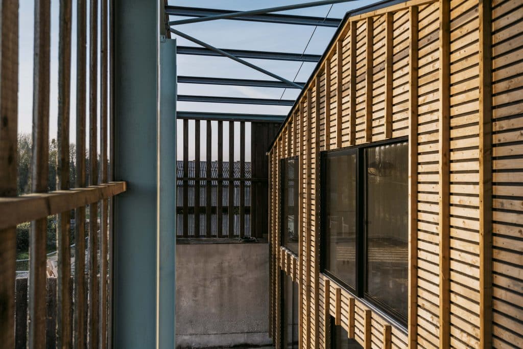 A modern eco barn conversion exterior with vertical wooden slats, large glass windows, and steel beams. Geometric lines and open sky highlight the blend of natural light and contemporary design elements.