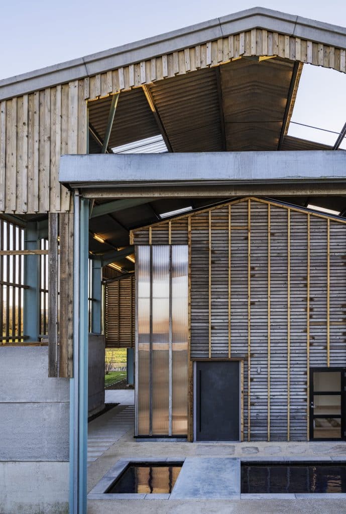 A modern barn-like structure features wooden slats, metal beams, and translucent panels. The roof peaks at the centre, and the facade has geometric lines and textures. A small reflective pool sits in the foreground, echoing the structure's clean design.