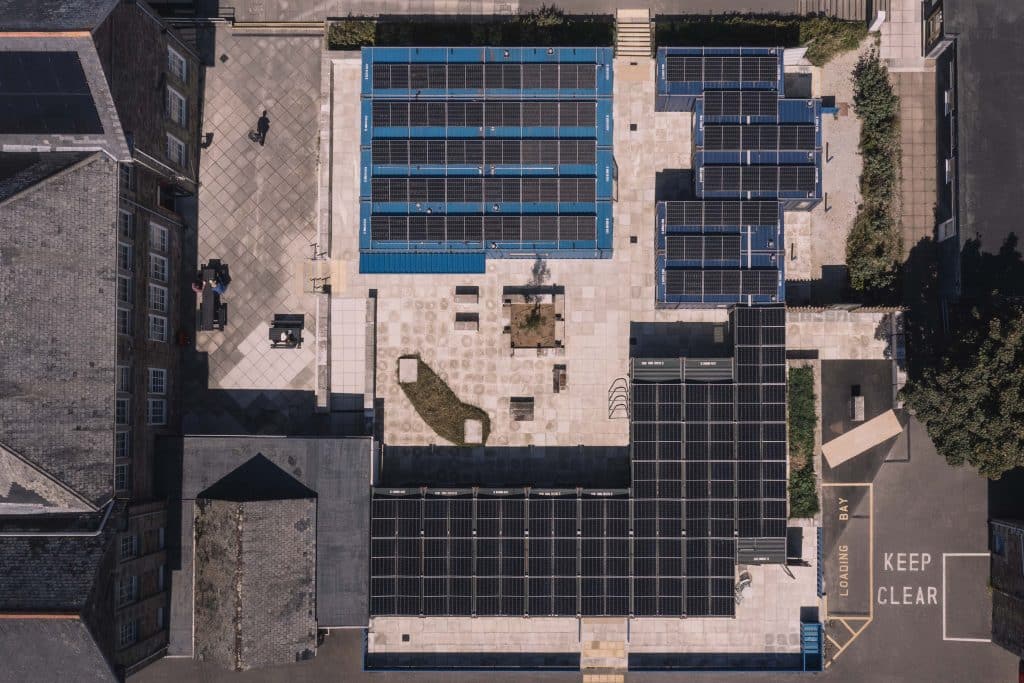 Aerial view of a courtyard with several building roofs covered in solar panels, paved walkways, benches, greenery, and a loading bay marked with KEEP CLEAR text on the ground.