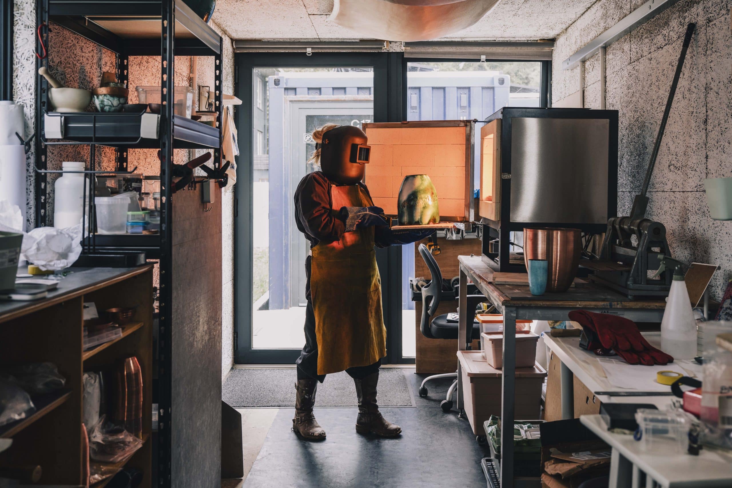 A person wearing protective gear, including a helmet and apron, stands in a workshop, carefully placing or removing an object from a glowing kiln. Shelves with tools and materials line the workspace.