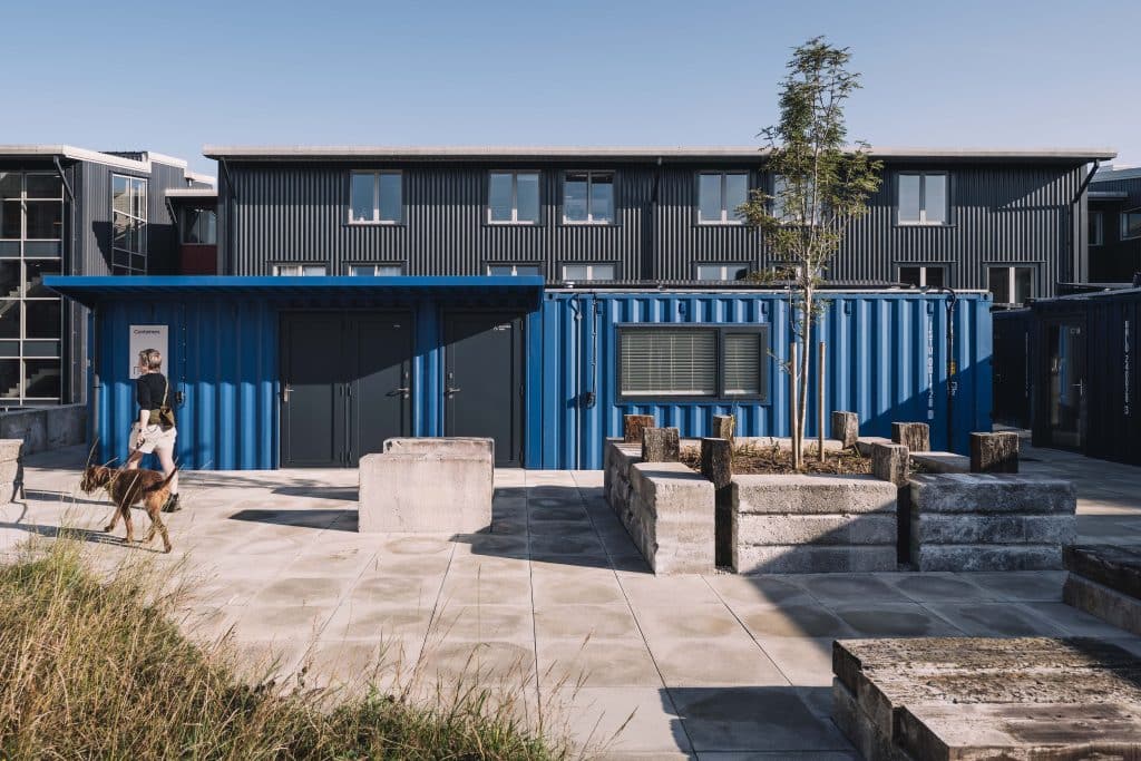 View through an open hallway with glass doors and wooden slats, leading to an outdoor area with stone planters and a young tree; a brick building with many windows is in the background.