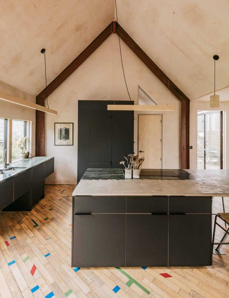 Modern kitchen with high vaulted ceiling, exposed wooden beams, and light wood floor with scattered colourful tile accents. Dark kitchen island with stone worktop sits centre, holding a small plant. Large windows and minimal decor create a bright, airy atmosphere.