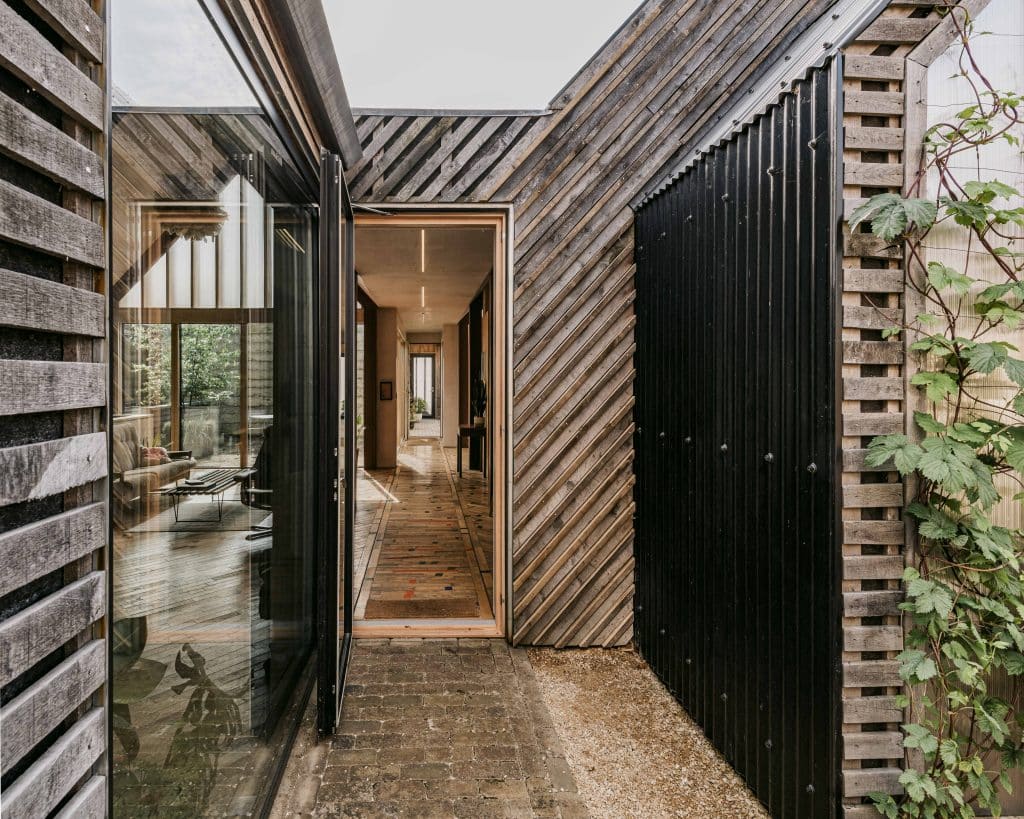 A narrow passageway with brick flooring and diagonal wooden cladding leads to a hallway inside a modern home. Large glass windows reflect the outdoor greenery, and plants climb a trellis beside the black panelled exterior wall. The interior appears bright and open.