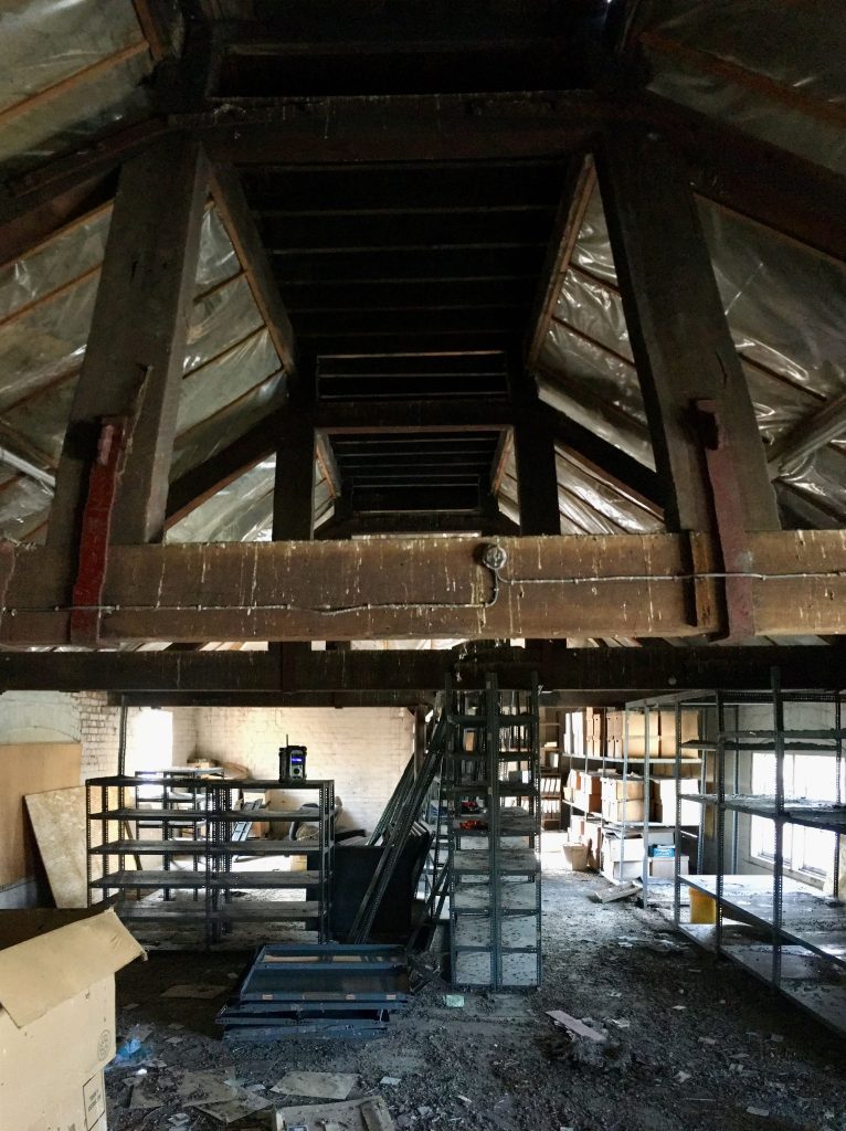 An attic-like space with exposed wooden beams and a steep central staircase. The area is cluttered with empty metal shelving units, boxes, and debris on the floor. The slanted ceiling is lined with metallic insulation panels, and natural light filters in.