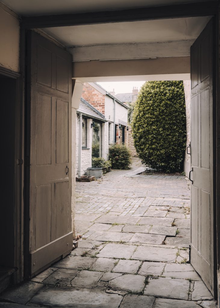 View through open double wooden doors showing a stone courtyard with uneven paving, potted plants, brick and stone buildings, and a tall, dense green bush at the centre right. The scene is quiet and appears to be in a historic area.