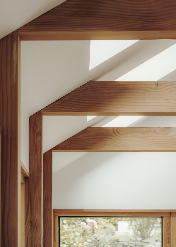 Sunlight streams through a white ceiling with exposed wooden beams in a modern interior, casting geometric shadows. Below, a window reveals blurred greenery outside. The overall atmosphere is bright, clean, and minimalist with natural wood accents.