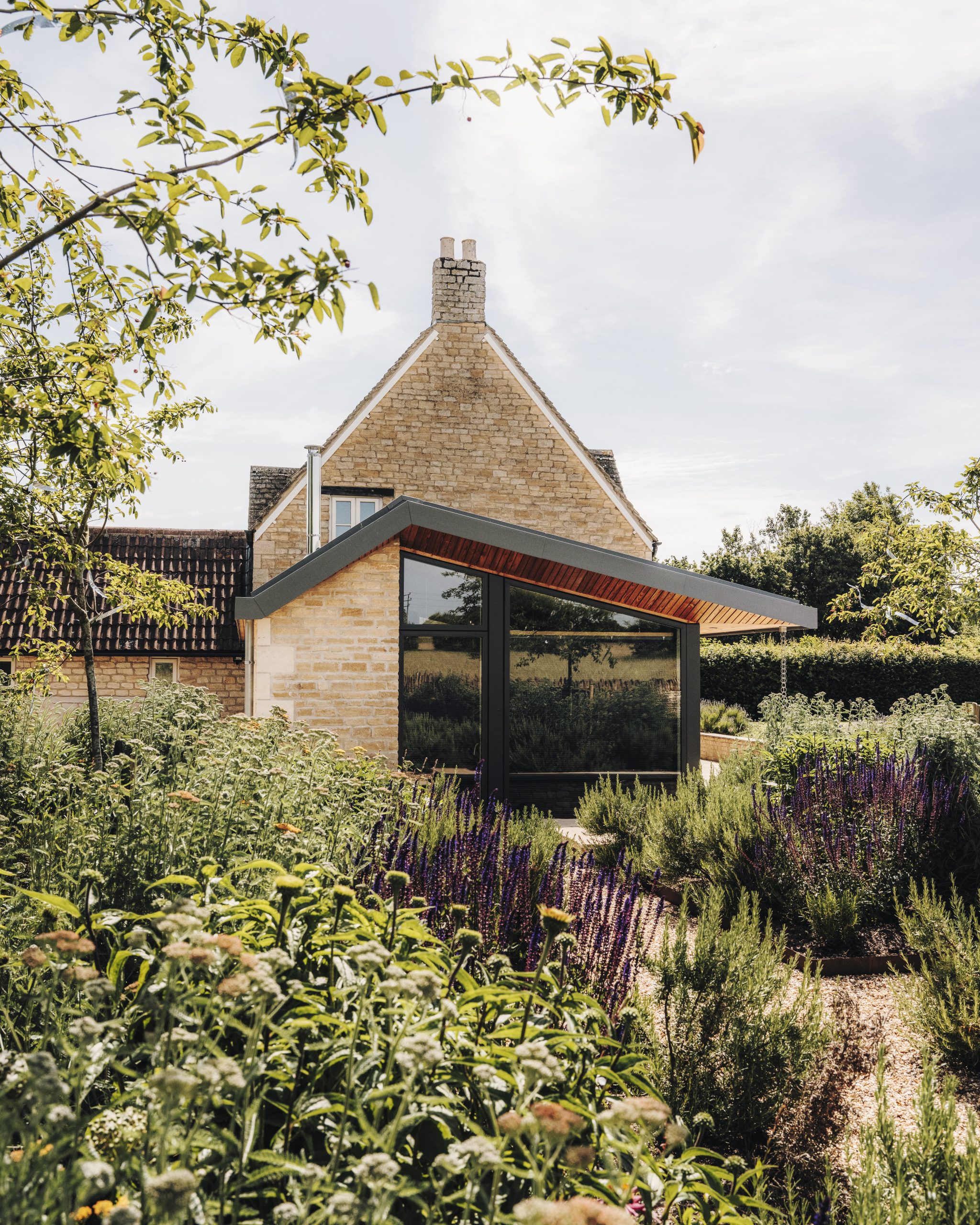 A modern glass extension with angled roof is attached to a traditional stone house, surrounded by lush green plants and purple flowers in a sunlit garden. The sky is clear and trees frame the peaceful scene.