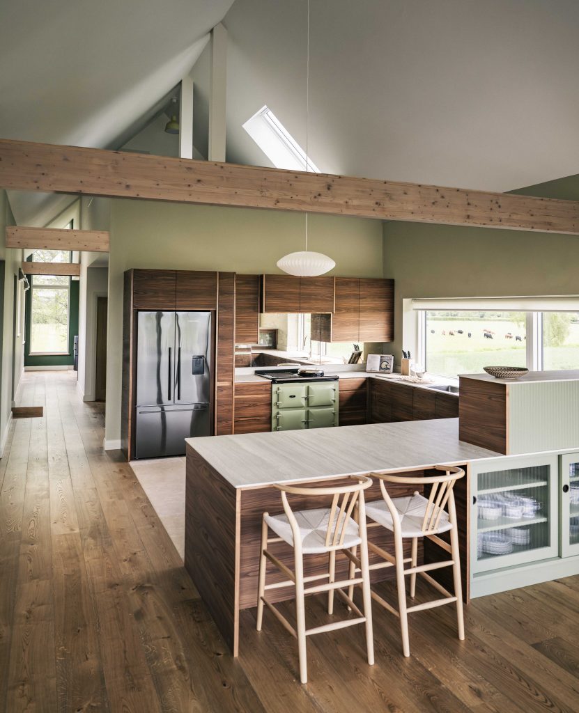 Modern kitchen with high vaulted ceiling, exposed timber beams, and sage green walls. Features wooden cupboards, stainless steel appliances, a pale island with two light wooden chairs, large windows with countryside views, and wooden plank flooring throughout.