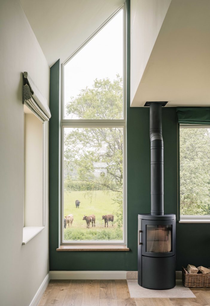 A modern living room with a tall window showing green fields and grazing cows outside. Inside, there is a black wood-burning stove with a vertical flue, green and cream walls, a woven basket with firewood, and wooden flooring.