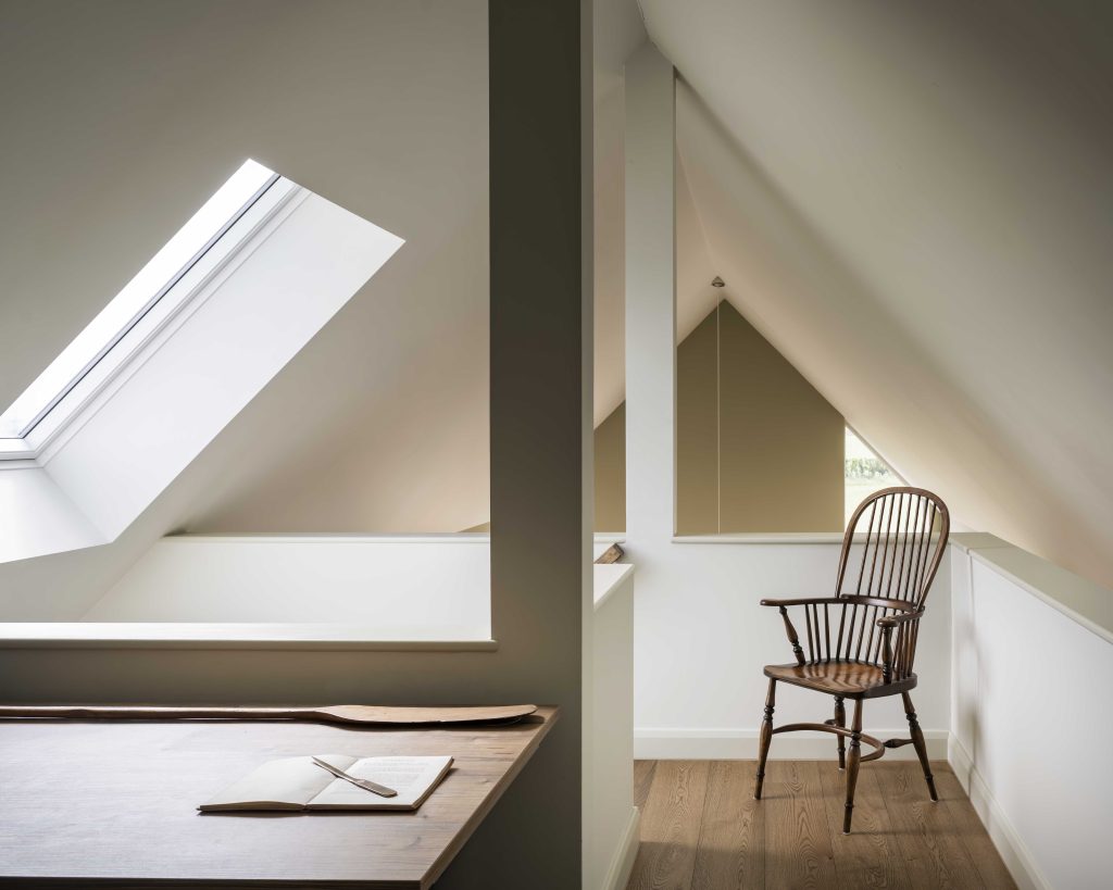 A bright attic room features slanted white ceilings with two skylights, a wooden desk with an open book, and a classic wooden chair on light wood flooring. Soft natural light fills the minimal, cosy space, highlighting its clean and serene aesthetic.