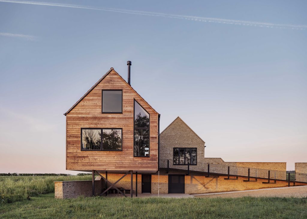 A modern house in a rural setting at sunset, featuring a raised wooden section with large geometric windows connected to a brick section. The building sits on stilts above grass, with a sloped ramp and tree reflections visible in the windows.