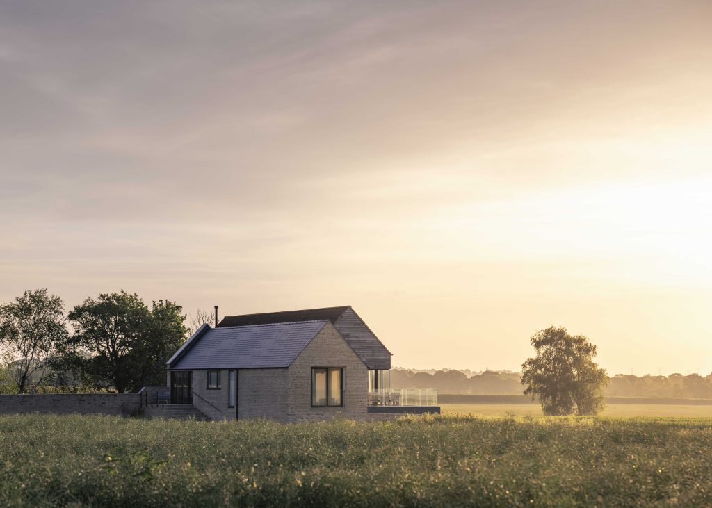 A small, modern house with a sloping roof stands alone in a green field at sunrise or sunset. Trees and a distant forest line the horizon under a soft, golden sky, bathing the landscape in warm light. The scene is calm and peaceful.