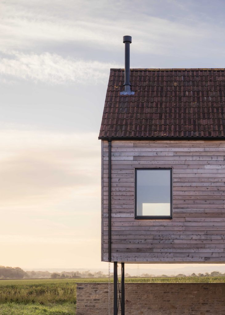 A modern wooden house on stilts is shown in the countryside at sunset. The house has a tiled roof, a chimney, a single square window, and timber panelling. Lush green fields and a cloudy sky stretch into the background.