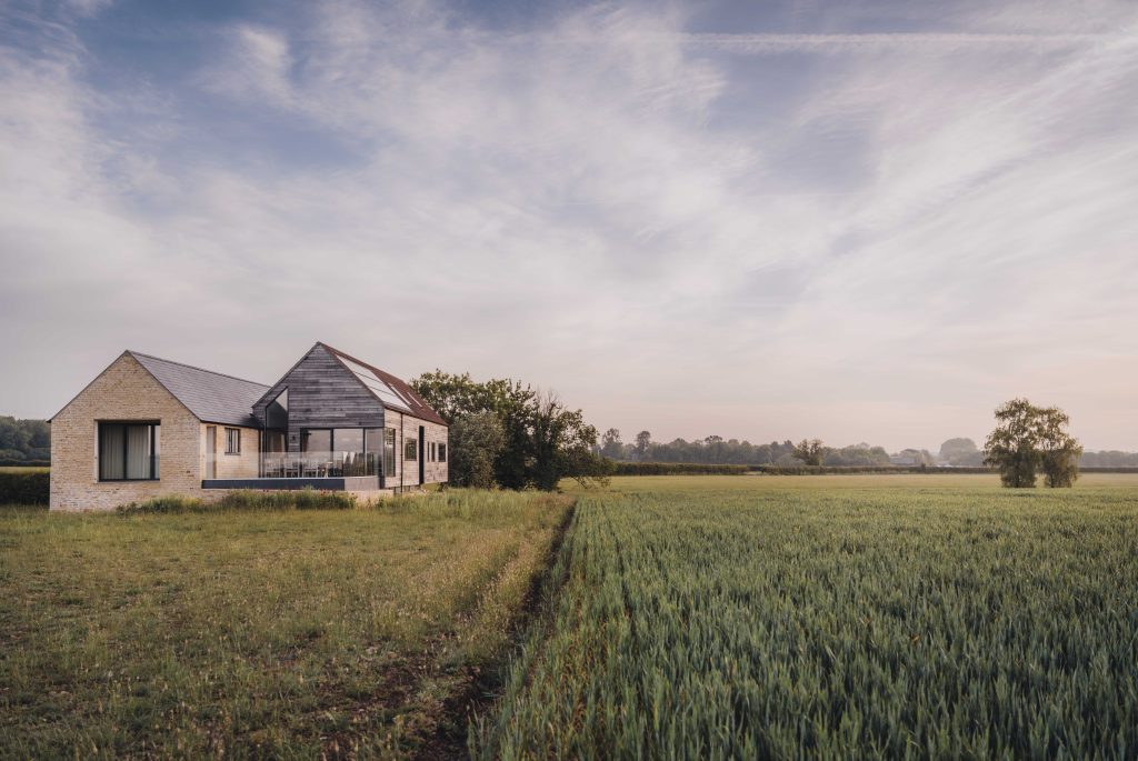 A modern house with stone and wood cladding stands on the edge of a green field under a cloudy sky. The left side of the house features large windows, and a covered veranda faces the open countryside. Trees and distant hedgerows border the field.